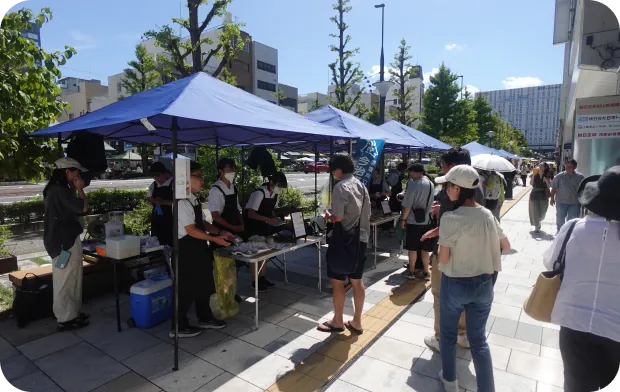 屋外の歩道に設置されたテント下で、スタッフが来場者に対応しているイベントの受付・出店の様子